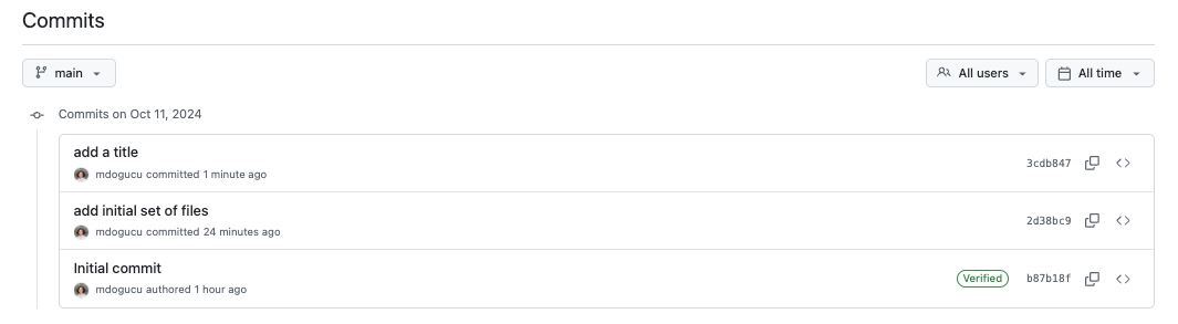Screenshot of a GitHub commit history page for the main branch. The page shows three commits made on October 11, 2024 by user mdogucu: 'add a title' (committed 1 minute ago), 'add initial set of files' (committed 24 minutes ago), and 'Initial commit' marked as Verified (authored 1 hour ago). The interface includes filter options for branch (main), users (All users), and time period (All time). Commit hashes 3cdb847, 2d38bc9, and b87b18f are visible.